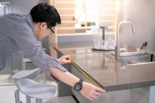 Genç Asyalı adam sergi salonundaki mutfak tezgahında granit tezgahları ölçmek için mezura kullanıyor. Ev dekorasyonu için mobilya alışverişi. İç tasarım kavramı