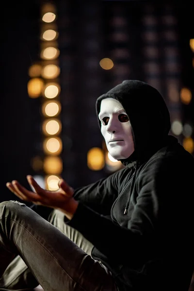 Mystery hoodie man in white mask sitting in the rain on rooftop of abandoned building at night. Bipolar disorder or Major depressive disorder. Depression concept