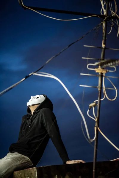 Mystery hoodie man in white mask sitting with antenna on rooftop of abandoned building looking up at the sky during the twilight time. Bipolar disorder or Major depressive disorder. Depression concept