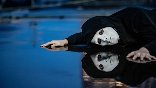 Reflection of mystery hoodie man in white mask feeling depressed and disappointed lying on wet floor after the rain. Sadness, depression and bipolar disorder concepts
