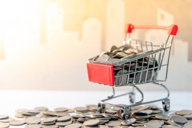 Saving money concept. Coins in mini shopping cart (trolley) and spilling on the table with city background.