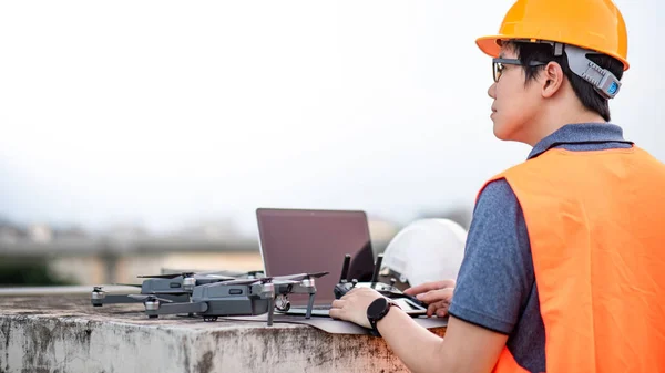 Asian engineer man working with drone, remote control and laptop computer at construction site. Using unmanned aerial vehicle (UAV) for land and building site survey in civil engineering project.