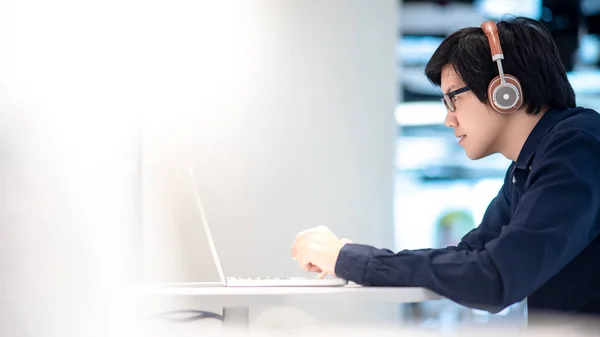 Asian business man listening to music by headphones while working with laptop computer in coworking space. Freelance or digital nomad lifestyle. Online shopping or E-commerce social media on internet