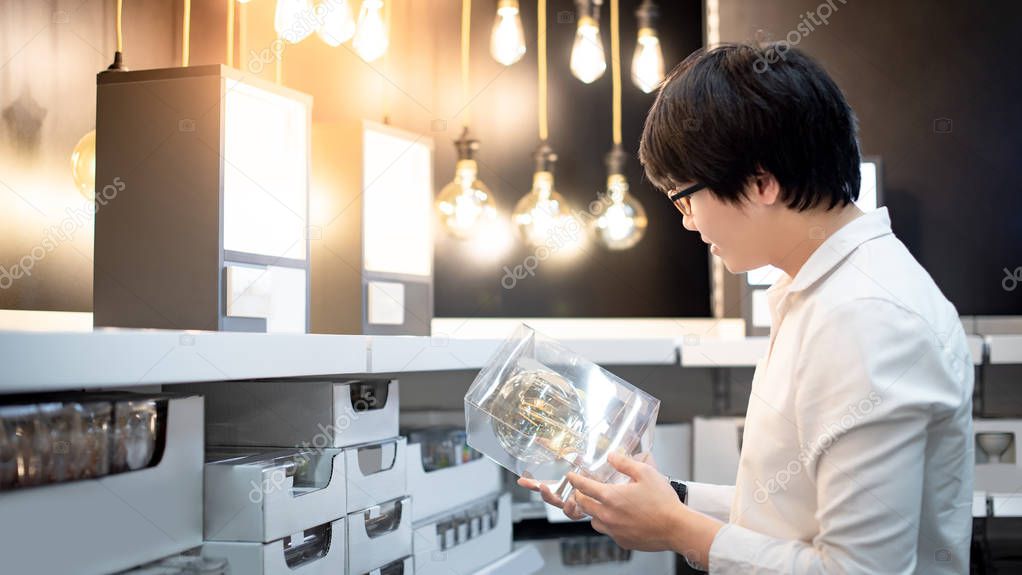 Asian guy shopper choosing package of electrical light bulb from product shelf in warehouse. Furniture home decoration shopping concept