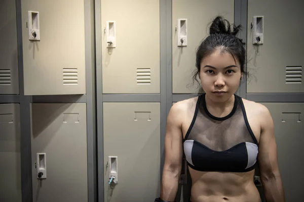 Confident Asian woman boxer with boxing gloves standing by the locker cabinet in locker room. Female boxing class concept