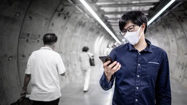Asyalı adam metro tünelinde akıllı telefon kullanarak kalabalık insanların yürüdüğü maske takıyor. Coronavirus (COVID-19) kamu alanındaki pandemik koruma. 2019-nCoV önleme kavramı