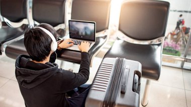 İş ve seyahat konsepti. Kapüşonlu Asyalı turist yolcu koltuğunda dizüstü bilgisayar kullanarak havaalanı terminalinin kalkış koridorunda uçuş beklerken kulaklıkla müzik dinliyor.