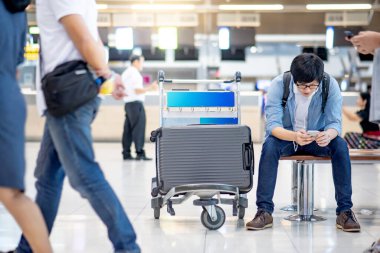 Tatil seyahati konsepti. Tramvayda valiziyle oturan Asyalı turist havaalanı terminalinin kalkış salonundaki bagaj kontrolünde akıllı telefonu havayolu için kullanıyor..