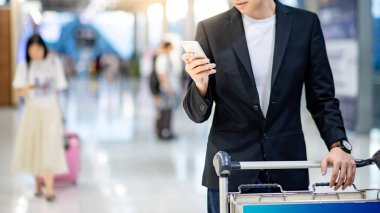 İş seyahati kavramı. Uluslararası havaalanı terminalinin kalkış salonunda tramvay arabasıyla yürürken resmi takım elbiseli akıllı Asyalı iş adamı havayolu çevrimiçi check-in için akıllı telefon kullanıyor..