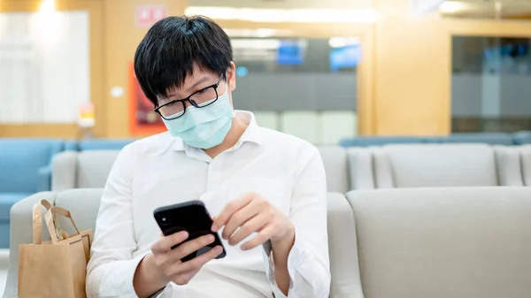 Male Asian patient wearing surgical face mask using smartphone at waiting area in hospital or medical center. Medical exam or body check up. Coronavirus pandemic prevention. Health care concept