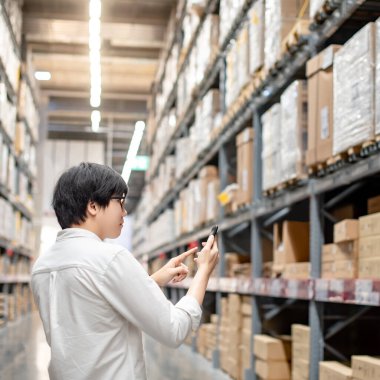Depo koridorunda durup akıllı telefondan alışveriş listesini kontrol eden Asyalı bir adam. Fabrika malları almak ya da satın almak. Alışverişkolik kavram