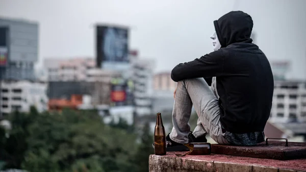 Mystery man in white mask with hoodie jacket sitting with broken alcoholic drink bottle on abandoned building feeling tired and stressed. Depression, mental health problem or alcoholism concept