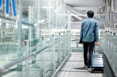 Havaalanı terminalinin girişinde yürüyen bavul ve dijital tablet taşıyan Asyalı turist. Seyahat sigortası kavramı