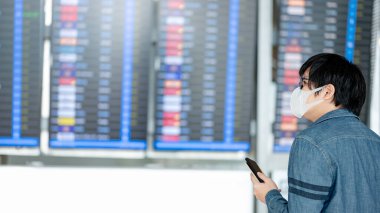 Yüz maskesi takan Asyalı turist havaalanı terminalinde akıllı telefon kullanarak varış panosundan kalkış biletini kontrol ediyor. Coronavirus (COVID-19) seyahat ederken salgını önler. Sosyal uzaklık kavramı