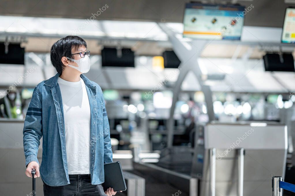 Asian man tourist wearing face mask carrying suitcase luggage at check-in counter in airport terminal. Coronavirus (COVID-19) pandemic prevention when travel. Health awareness and social distancing
