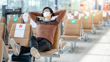 Korumalı yüz maskesi takan Asyalı turist havaalanı terminalinde valiziyle oturuyor. Coronavirus (COVID-19) yurtdışına seyahat ederken önleme. Sağlık farkındalığı ve sosyal uzaklık