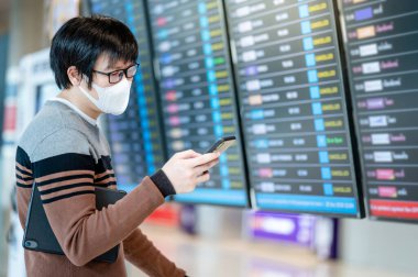 Yüz maskesi takan Asyalı turist havaalanı terminalinde akıllı telefon kullanarak varış panosundan kalkış biletini kontrol ediyor. Coronavirus (COVID-19) seyahat ederken salgını önler. Sosyal uzaklık kavramı