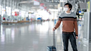 Yüz maskesi takan Asyalı turist havaalanı terminalinde kayıt için bavul taşıyor. Coronavirus (COVID-19) seyahat ederken salgını önler. Sağlık farkındalığı ve sosyal uzaklık
