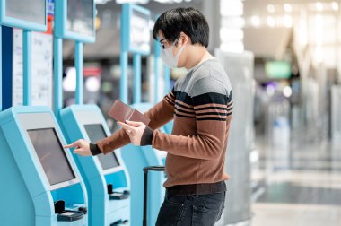 Asyalı turist yüz maskesi takıyor havaalanı terminalinde kendini kontrol gişesi kullanıyor. Yurtdışına seyahat ederken koronavirüs (COVID-19) salgını önleme. Sağlık farkındalığı ve sosyal uzaklık kavramı
