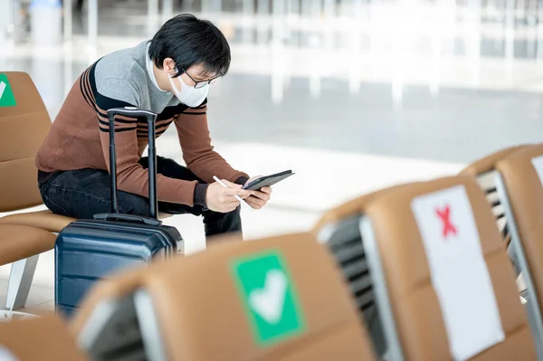 Asian man tourist with suitcase luggage wearing face mask using digital tablet in airport terminal. Coronavirus (COVID-19) prevention when travel abroad. Health awareness and social distancing concept