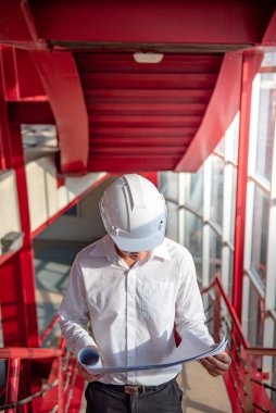 Asyalı inşaat mühendisi ya da koruma kaskı takan inşaat işçisi. Elinde planlar var. Erkek mimar inşaat alanında kırmızı merdivende çalışıyor. İnşaat ve mimari tasarım kavramları