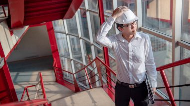 Asyalı inşaat mühendisi ya da miğfer takan inşaat işçisi elinde belge ve dizüstü bilgisayar tutuyor. Erkek mimar inşaat alanında kırmızı merdivende çalışıyor. Mimarlık konsepti