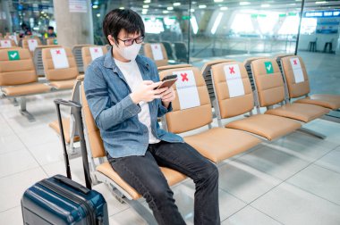 Çantalı Asyalı turist havaalanı terminalinde akıllı telefon kullanarak maske takıyor. Yurtdışına seyahat ederken koronavirüs (COVID-19) salgını önleme. Sağlık farkındalığı ve sosyal uzaklık