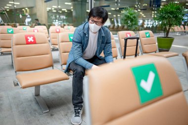 Korumalı yüz maskesi takan Asyalı turist havaalanı terminalinde bavuluyla oturuyor. Yurtdışına seyahat ederken koronavirüs (COVID-19) salgını önleme. Sağlık farkındalığı ve sosyal uzaklık