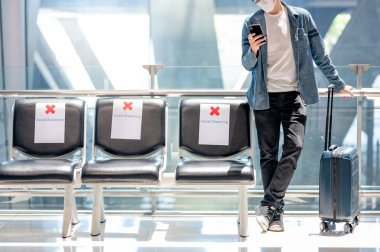 Çantalı Asyalı turist havaalanı terminalinde akıllı telefon kullanarak maske takıyor. Yurtdışına seyahat ederken koronavirüs (COVID-19) salgını önleme. Sağlık farkındalığı ve sosyal uzaklık