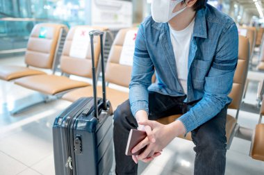 Korumalı yüz maskesi takan Asyalı turist havaalanı terminalinde bavuluyla oturuyor. Yurtdışına seyahat ederken koronavirüs (COVID-19) salgını önleme. Sağlık farkındalığı ve sosyal uzaklık