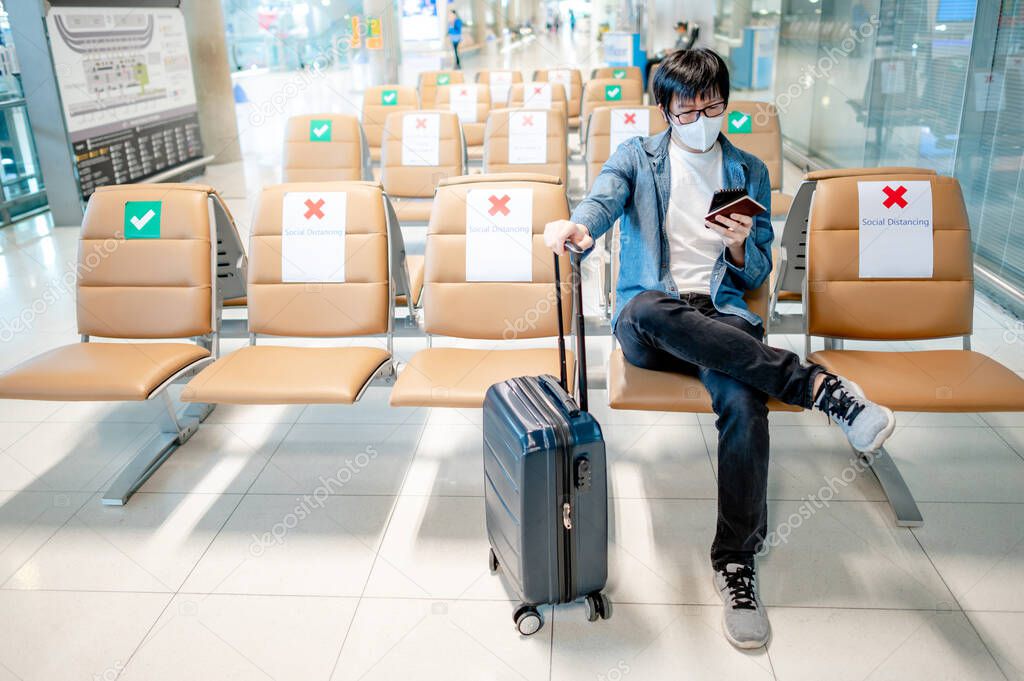 Asian man tourist with suitcase luggage wearing face mask using smartphone in airport terminal. Coronavirus (COVID-19) pandemic prevention when travel abroad. Health awareness and social distancing