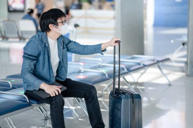 Havaalanı terminalinde yüz maskesi takan Asyalı bir turist dijital tablet ve bavul taşıyor. Coronavirus (COVID-19) yurtdışına seyahat ederken önleme. Sağlık farkındalığı ve sosyal uzaklık