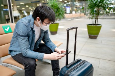 Çantalı Asyalı turist havaalanı terminalinde akıllı telefon kullanarak maske takıyor. Yurtdışına seyahat ederken koronavirüs (COVID-19) salgını önleme. Sağlık farkındalığı ve sosyal uzaklık