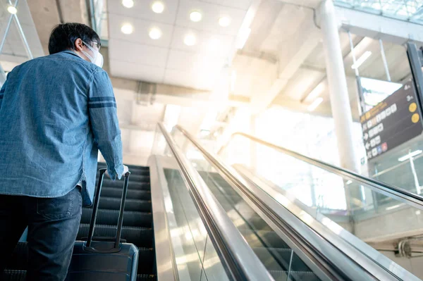 Travel insurance concept. Asian man tourist with face mask carrying suitcase luggage on escalator in airport terminal. Coronavirus (COVID-19) pandemic prevention when travelling.