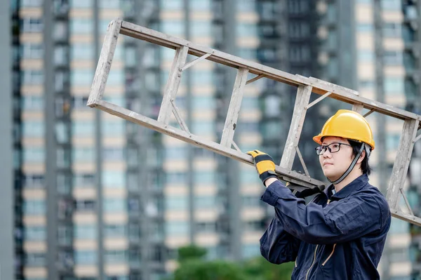 Asian maintenance worker man with protective suit and safety helmet carrying aluminium step ladder at construction site. Civil engineering, Architecture builder and building service concepts