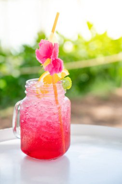 Pembe çiçekli soğuk kırmızı limonlu soda ve kumsaldaki kafede cam kavanozda limon dilimi. Taze yaz kokteyl içeceği konsepti