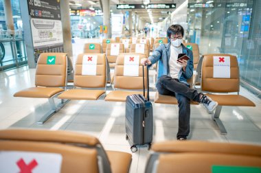 Çantalı Asyalı turist havaalanı terminalinde akıllı telefon kullanarak maske takıyor. Yurtdışına seyahat ederken koronavirüs (COVID-19) salgını önleme. Sağlık farkındalığı ve sosyal uzaklık
