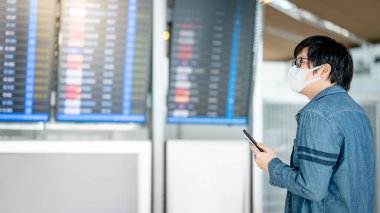 Yüz maskesi takan Asyalı turist havaalanı terminalinde akıllı telefon kullanarak varış panosundan kalkış biletini kontrol ediyor. Coronavirus (COVID-19) seyahat ederken salgını önler. Sosyal uzaklık kavramı