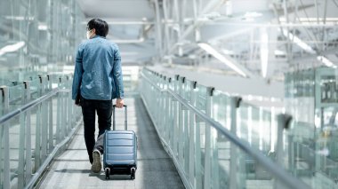 Yüz maskesi takan Asyalı turist havaalanı terminalinin kapısında bavul taşıyordu. Coronavirus (COVID-19) seyahat ederken salgını önler. Sağlık farkındalığı ve sosyal uzaklık