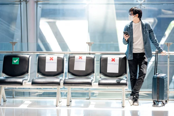 Asian man tourist with suitcase luggage wearing face mask using smartphone in airport terminal. Coronavirus (COVID-19) pandemic prevention when travel abroad. Health awareness and social distancing