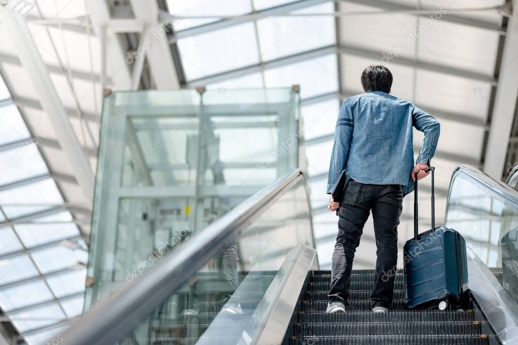 Travel insurance concept. Asian man tourist carrying suitcase luggage and digital tablet on escalator in airport terminal.