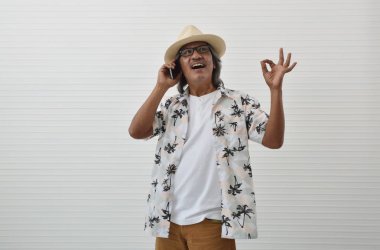 Hasır şapka, Gözlük, Yaz gömlek ve modern akıllı telefonda konuşurken ve ok parmak gösterilen beyaz duvar arka plan iş yaz tatil kavramı üzerinde oturum kahverengi şort giyen mutlu üst düzey traveler Asya adam