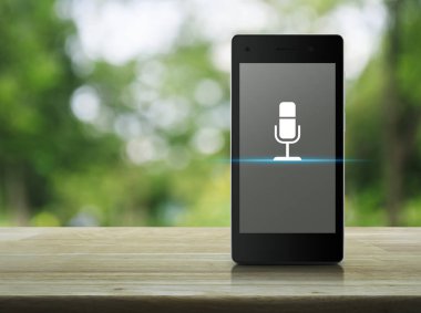 Modern akıllı cep telefonu ekranındaki mikrofon yassı simgesi parktaki bulanık yeşil ağacın üzerindeki ahşap masa üzerinde iş iletişimi online konsepti
