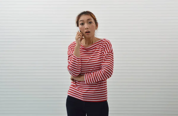 Confused young asian woman wearing red and white stripped shirt and jeans talking on modern smart phone over white wall background, Confuse facial expression