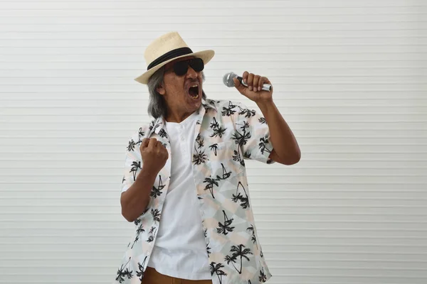 Funny senior traveler asian man wearing straw hat, sunglasses and summer shirt singing with microphone standing over white wall background, Business summer holiday concept