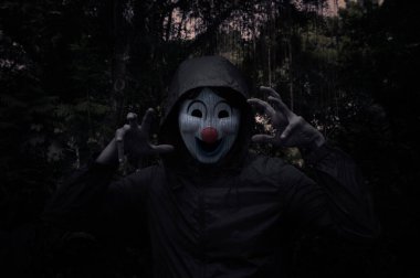 Korkunç kötü palyaço ceketi ağaçlı, yapraklı ve sarmaşıklı ürkütücü karanlık ormanın üzerinde duruyor, Cadılar Bayramı gizemi kavramı.