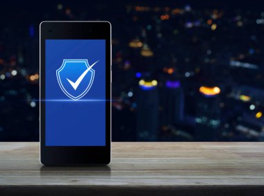 Güvenlik kalkanı, modern akıllı cep telefonu ekranında düz simge ile ahşap masa üzerinde bulanık gece lambası şehir kulesi ve gökdelen, Teknoloji İnternet siber güvenliği online konsepti