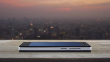 Modern akıllı cep telefonu ekranında fotokopi alanı olan ev simgesi ahşap masa üzerinde sıcak gün batımında şehir manzarası, İş dünyası emlak online konsepti