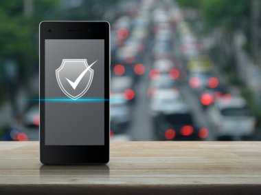 Güvenlik kalkanı, modern akıllı cep telefonu ekranında yassı bir simgeye sahip. Ahşap masanın üstünde, trafiğin yoğun olduğu saatte, şehirde arabalar ve yollar ile birlikte. Teknoloji internet siber güvenliği online konsepti.