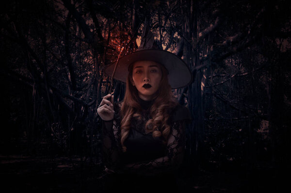 Halloween witch holding magic wand standing over spooky dark forest with tree, leaves and vine, Halloween mystery concept